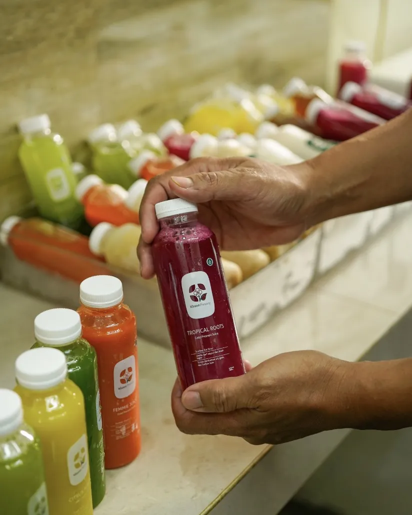 Tangan memegang sebotol jus merah Kboon dengan label isi dan cara simpan